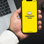 Mão segurando um smartphone com o aplicativo do Mercado Livre aberto na tela amarela, em uma mesa de trabalho com um notebook prateado e objetos de escritório ao fundo.