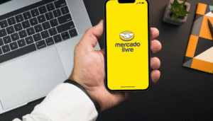Mão segurando um smartphone com o aplicativo do Mercado Livre aberto na tela amarela, em uma mesa de trabalho com um notebook prateado e objetos de escritório ao fundo.