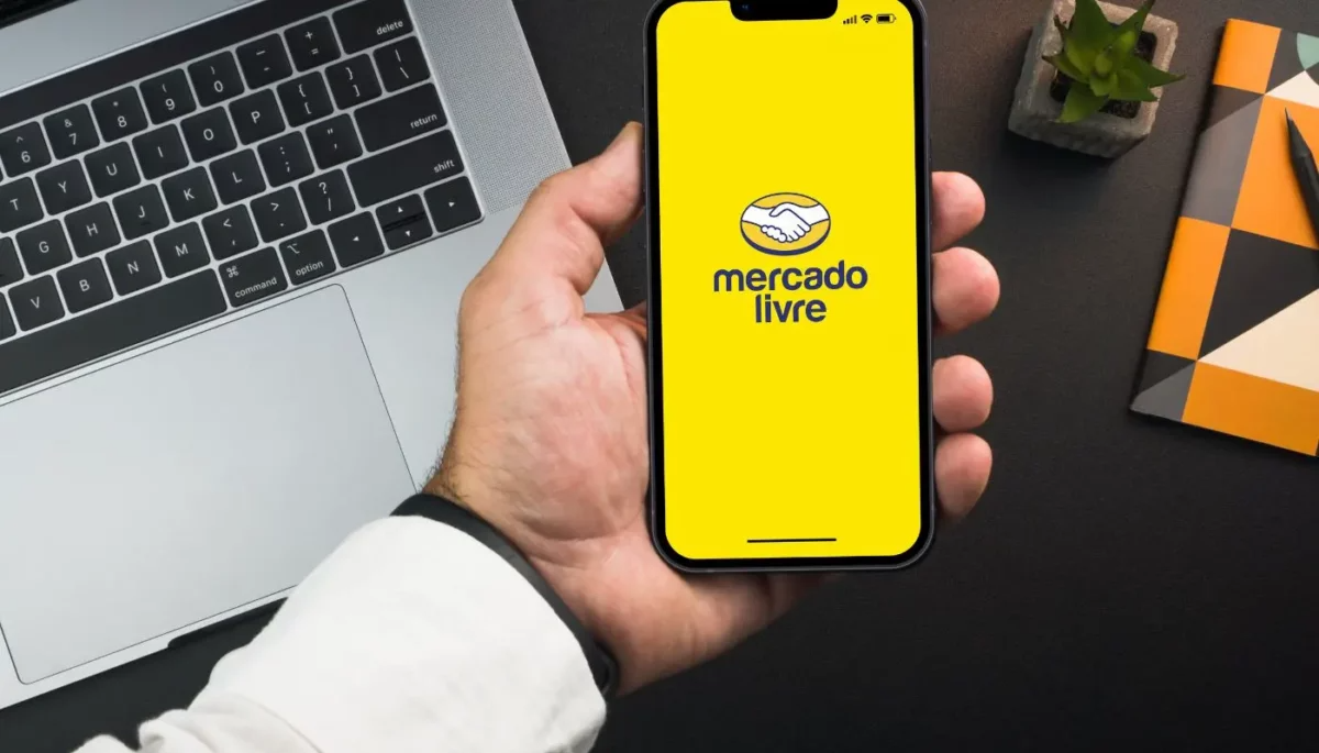 Mão segurando um smartphone com o aplicativo do Mercado Livre aberto na tela amarela, em uma mesa de trabalho com um notebook prateado e objetos de escritório ao fundo.